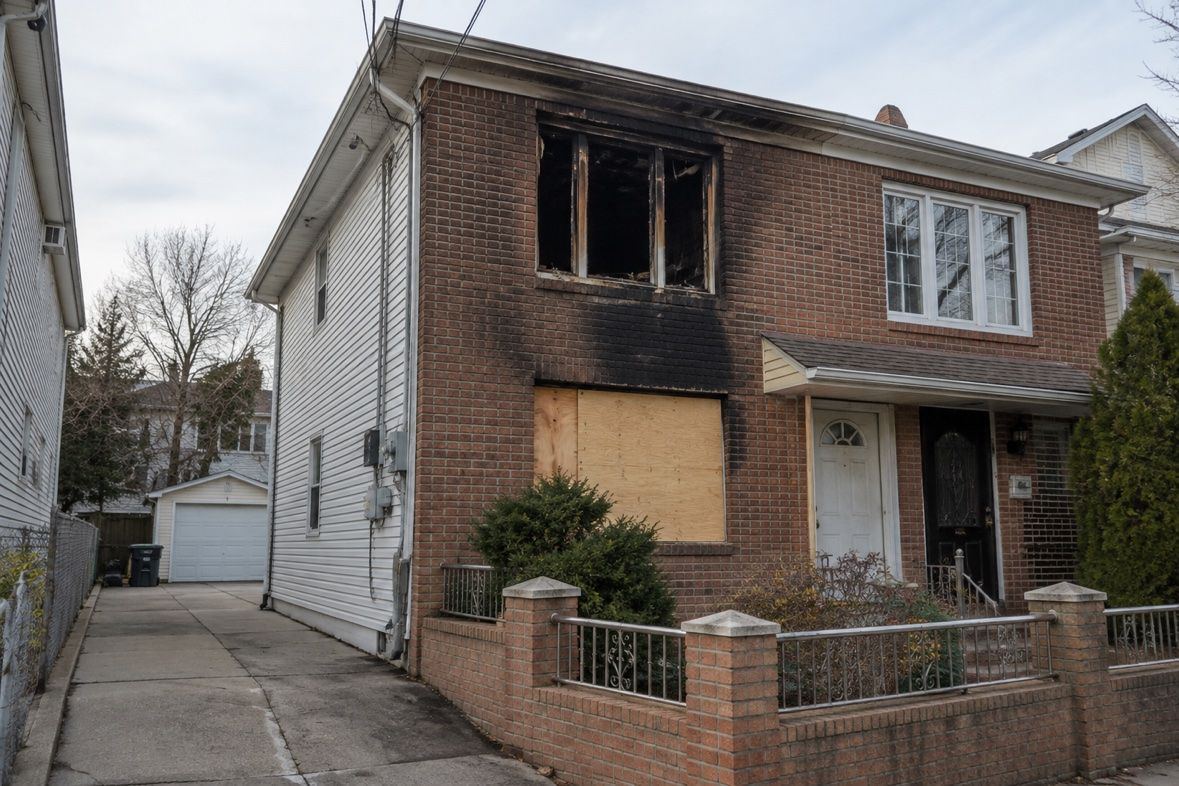 Brick house with a burned-out upper window, soot-stained brickwork and a boarded lower opening.